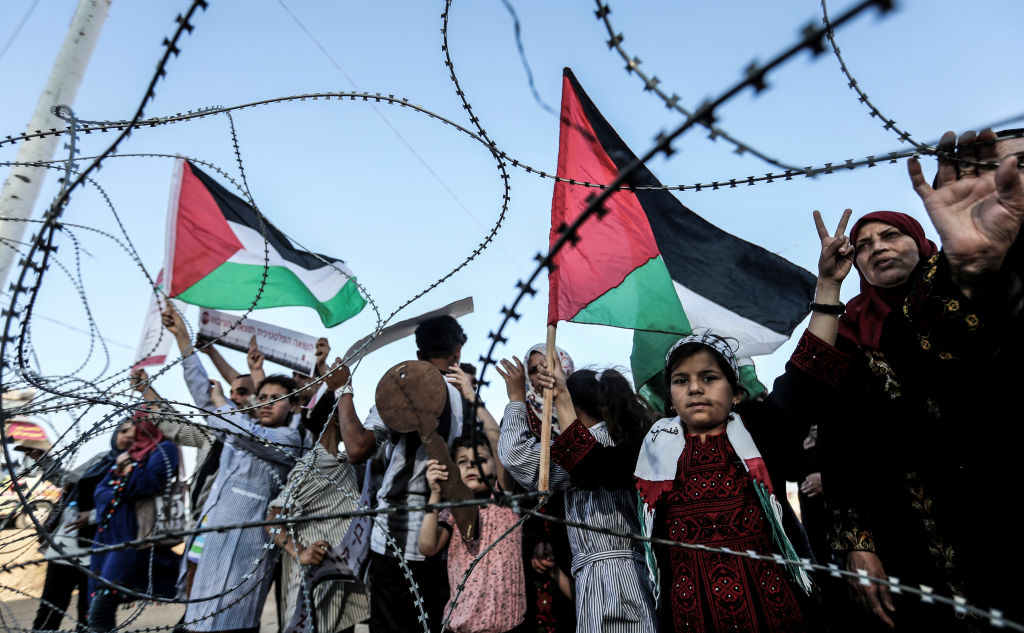 A girl raises a Palestinian flag as another Palestinian boy holds a wooden key symbolising the return, as they stand with others before the barbed-wire marking the border between the Gaza strip and Israel east of Gaza City on May 13, 2018 during a demonstration anticipating the 70 years since the Palestinian "Nakba", or catastrophe, of 1948 when over 700,000 Palestinians fled or were expelled from their homes in the 1948 war surrounding Israel's creation. (Photo by MAHMUD HAMS / AFP) (Photo credit should read MAHMUD HAMS/AFP via Getty Images)