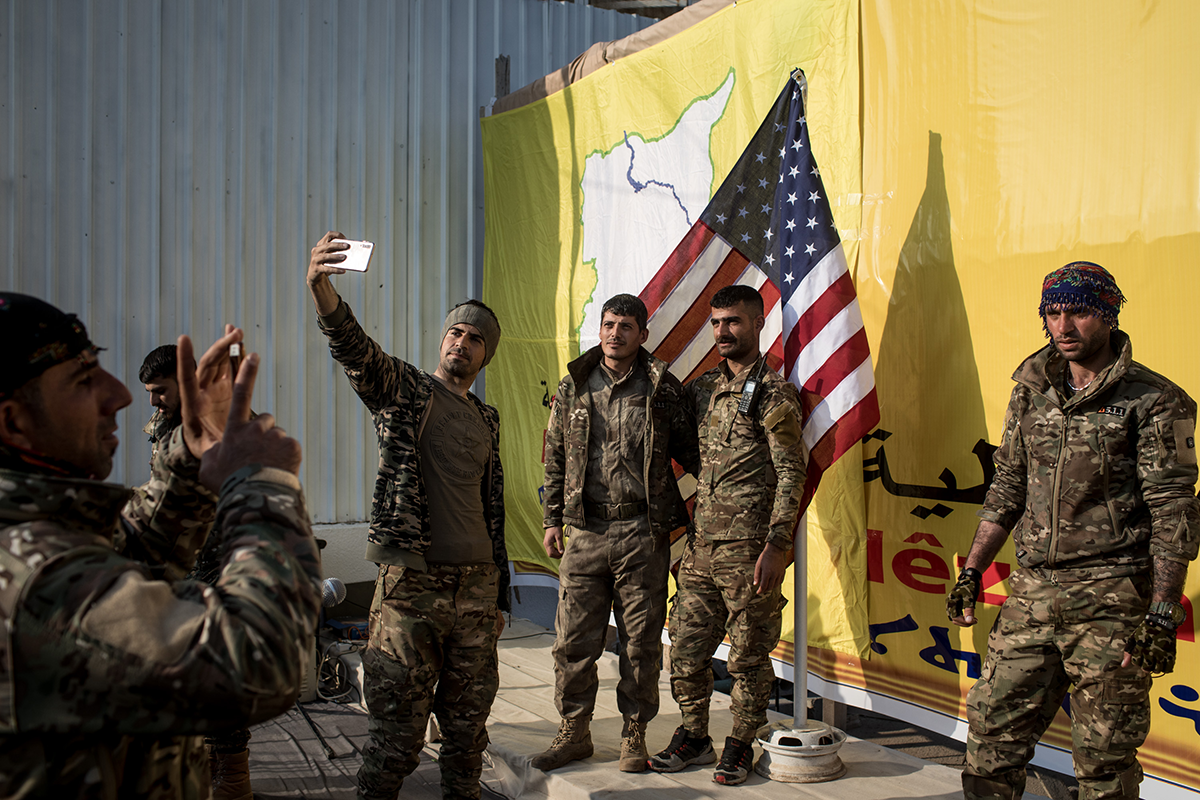 SDF fighters pose for a photo with the American flag on stage after a SDF victory ceremony announcing the defeat of ISIL in Baghouz was held at Omer Oil Field on March 23, 2019 in Baghouz, Syria. (Photo by Chris McGrath/Getty Images.)