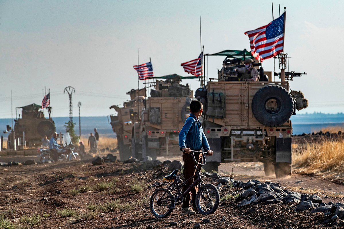 A Syrian boy on his bicycle looks at a convoy of US armored vehicles patrolling fields near the northeastern town of Qahtaniyah at the border with Turkey, on October 31, 2019. (Photo by DELIL SOULEIMAN/AFP via Getty Images.)
