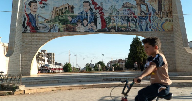 A young boy rides his bicycle in the southern Syrian city of Daraa on August 14, 2018. Behind him is a gate ornated with images of Syrian President Bashar al-Assad (L) and his late father Hafez al-Assad. (Photo by Andrei BORODULIN / AFP) (Photo credit should read ANDREI BORODULIN/AFP/Getty Images)