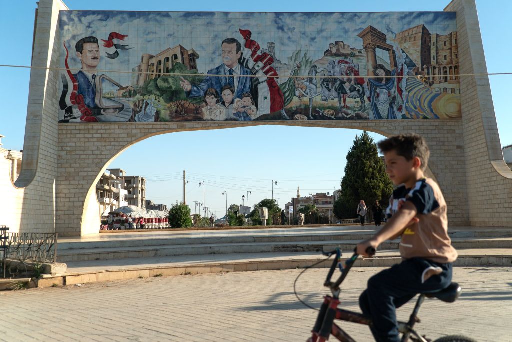 A young boy rides his bicycle in the southern Syrian city of Daraa on August 14, 2018. Behind him is a gate ornated with images of Syrian President Bashar al-Assad (L) and his late father Hafez al-Assad. (Photo by Andrei BORODULIN / AFP) (Photo credit should read ANDREI BORODULIN/AFP/Getty Images)