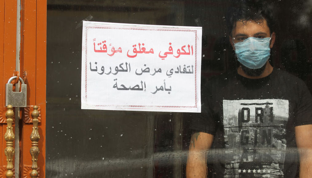 An Iraqi man, wearing a protective mask, stands inside a coffee shop with a sign in Arabic which reads 