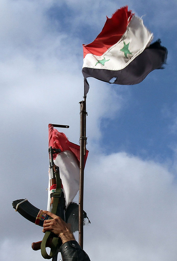 A Syrian soldier lifts up his machine gun next to the Syrian flag, in the southern city of Sweida on January 23, 2013. The two provinces are diametrically opposed in the current conflict. If Daraa chose rebellion, Sweida is firmly in regime hands. AFP PHOTO/ANWAR AMRO (Photo credit should read ANWAR AMRO/AFP via Getty Images)